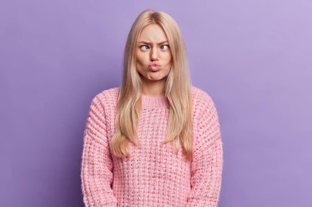 Funny blonde young woman has silly grimace crosses eyes and pouts lips makes brainless face wears kn